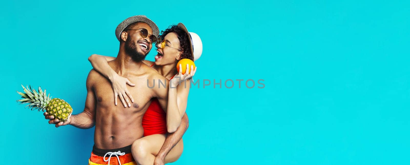A young couple in swimwear poses in front of a blue background. The man is holding a pineapple and the woman is holding an orange. They are both smiling and looking at each other.