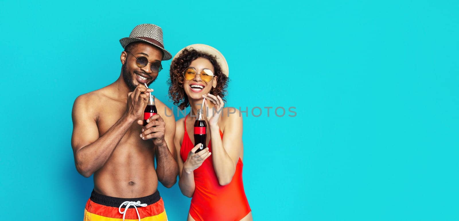 A young black couple dressed in swimwear and sunglasses smiles and enjoys drinks together on a bright blue background.