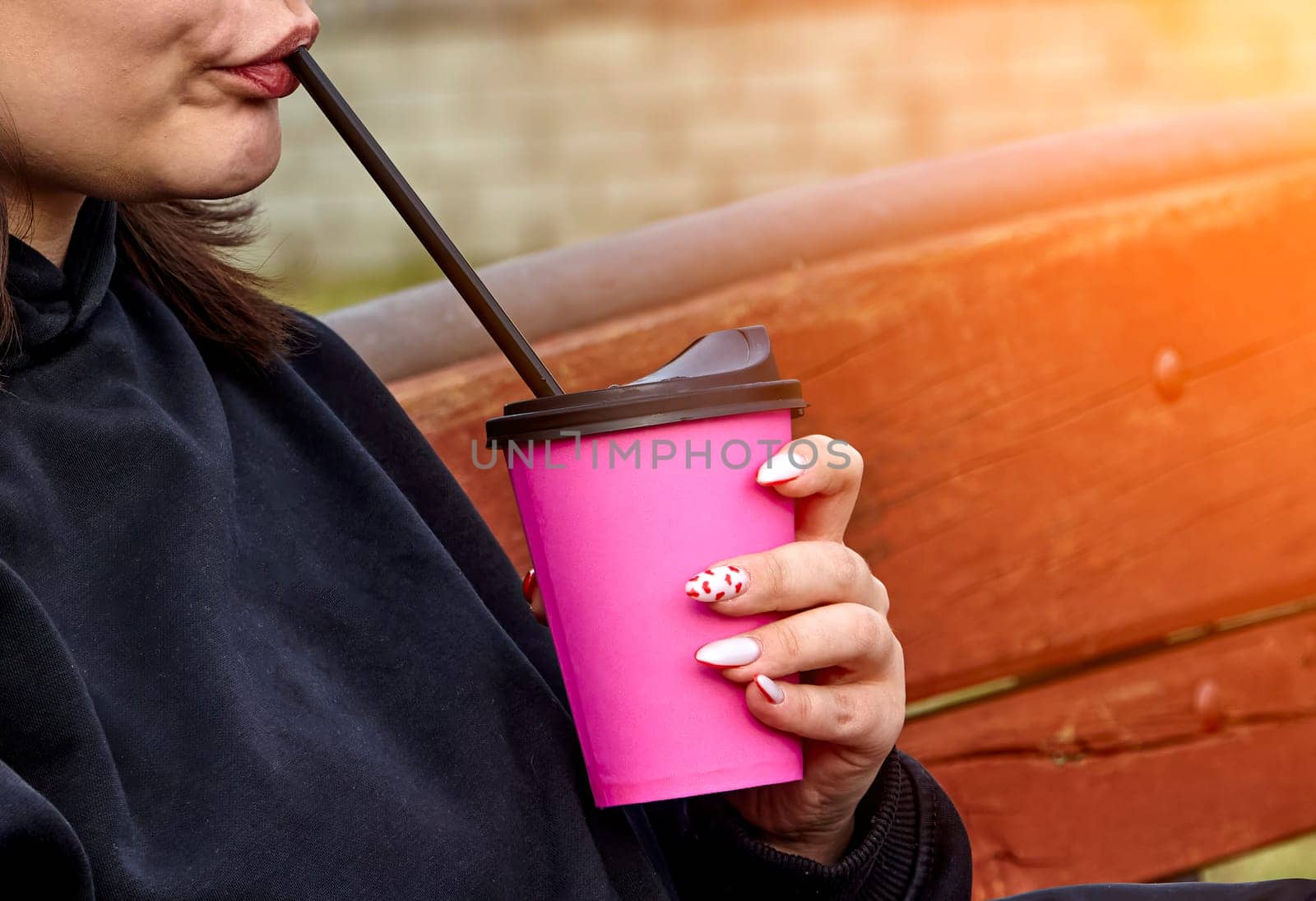 A young woman with long brown hair and white headphones is drinking from a pink cup with a straw. She is wearing a black sweatshirt and has her nails painted pink with white polka dots.