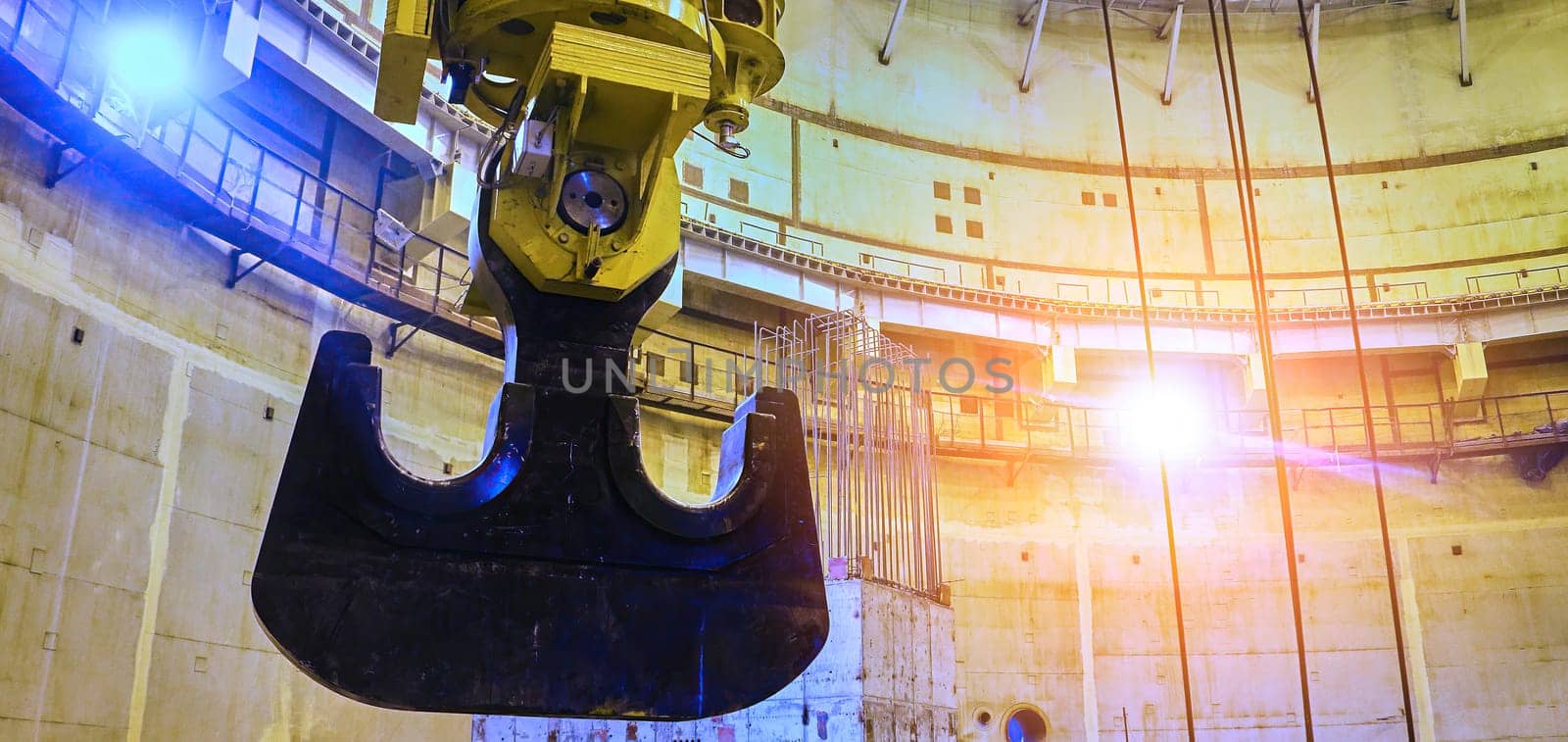 A large, steel claw hangs suspended from a crane inside a vast, concrete structure. The claw appears to be reaching towards a shaft of sunlight that illuminates the room. The structure appears to be under construction and is surrounded by scaffolding and other building materials.