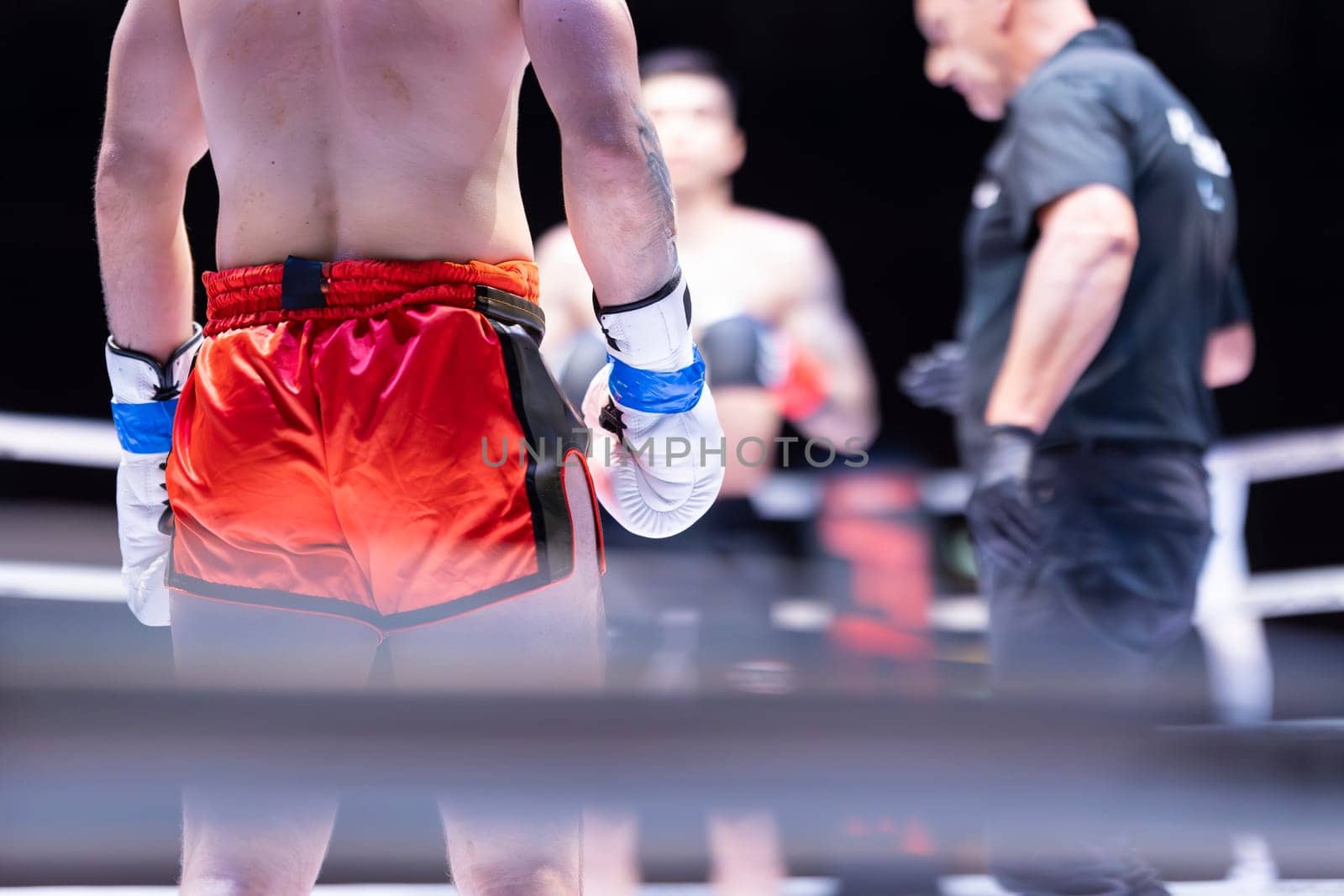 A man in a red boxing shorts is standing in a ring with his hands on his hips. A referee is standing behind him