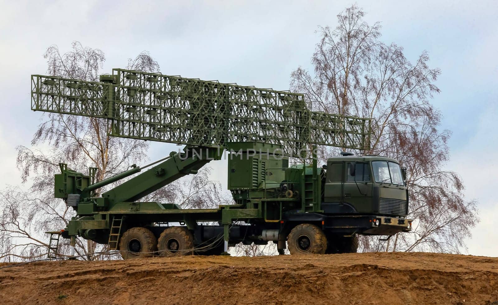 A large, military radar system is mounted on a truck and positioned on a dirt hilltop. The radar dish is extended and pointed upwards, likely scanning for potential threats in the sky.