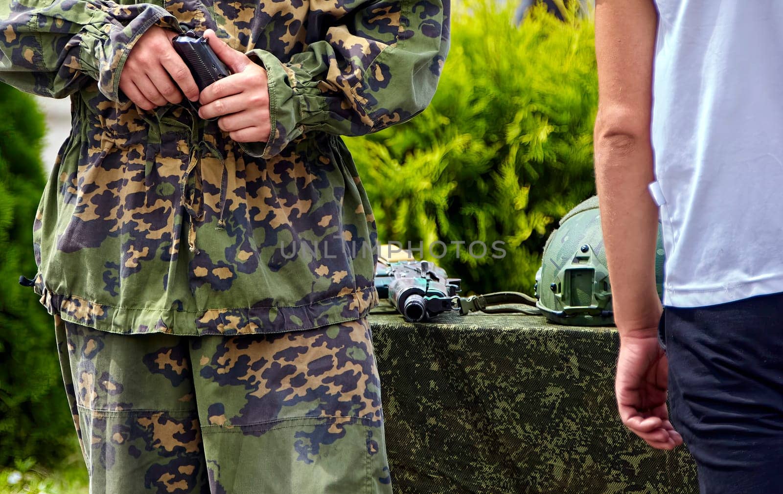 A soldier in full camouflage gear stands with a pistol in his hand, while a civilian walks away in the background. The soldier is focused on his task, while the civilian seems to be unconcerned about the situation. They are surrounded by greenery, suggesting a training exercise or a military event.