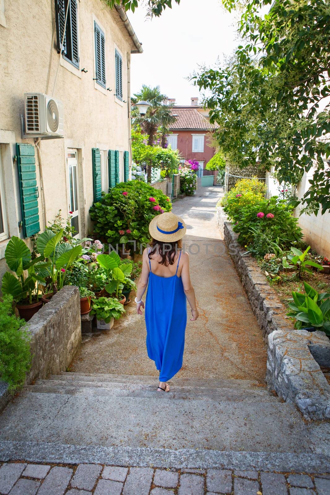 A woman in a blue dress and straw hat walks down a quaint alley lined with green plants and flowers