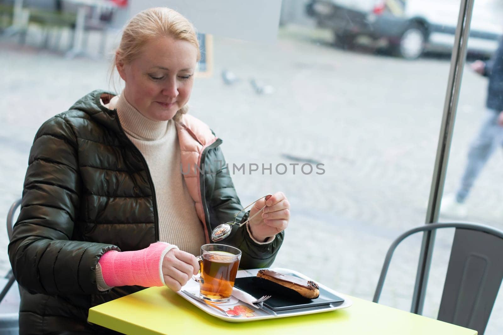 woman with a pink plaster cast on her right arm sits in a cafe and drinks tea with eclair, active life despite restrictions