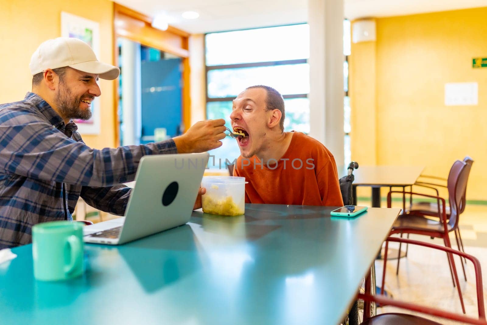 Caucasian male colleague feeding a man with disability in a coworker cafeteria