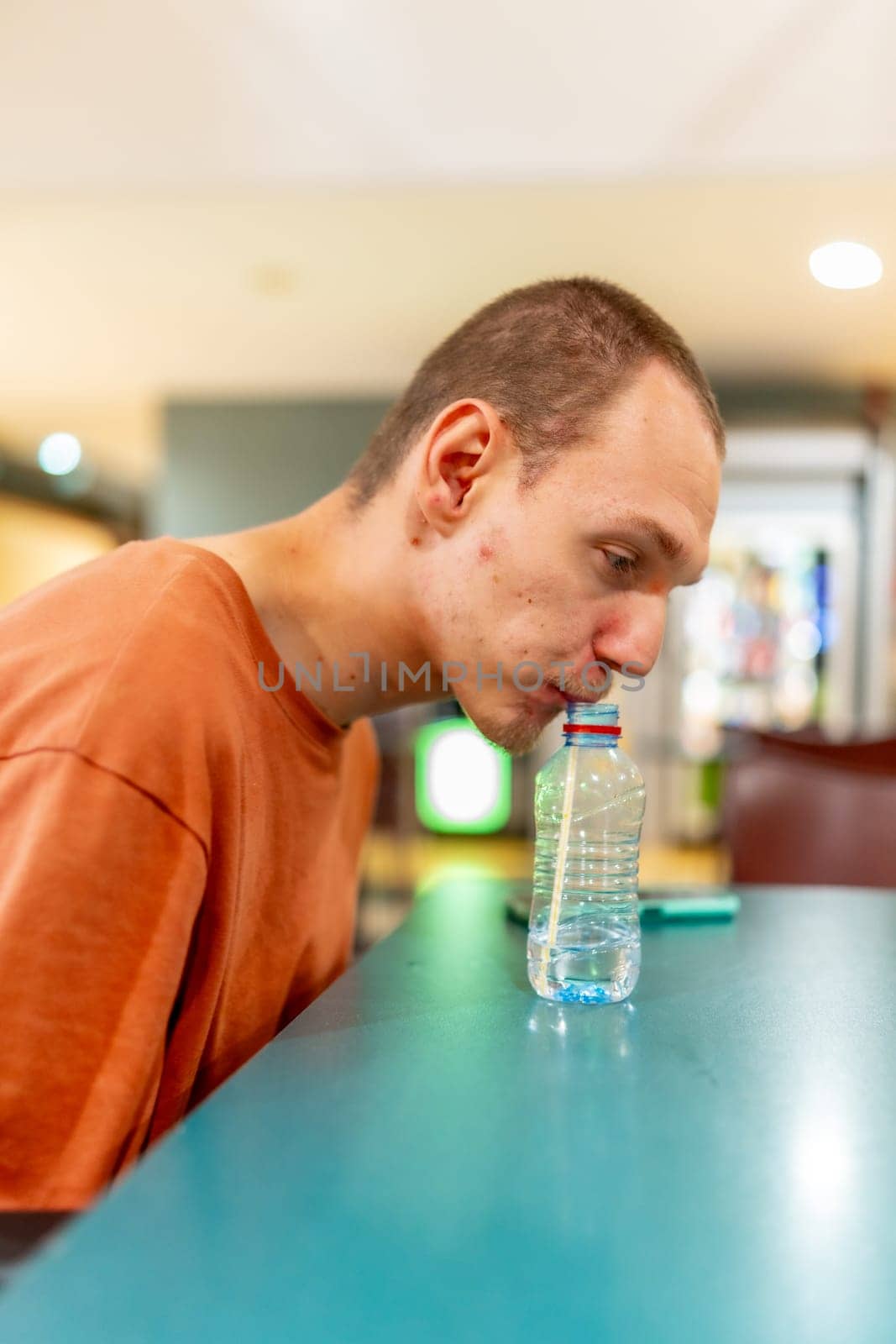 Vertical photo of a caucasian adult man with cerebral palsy achieve to drink water helped by straw in a coworking cafeteria