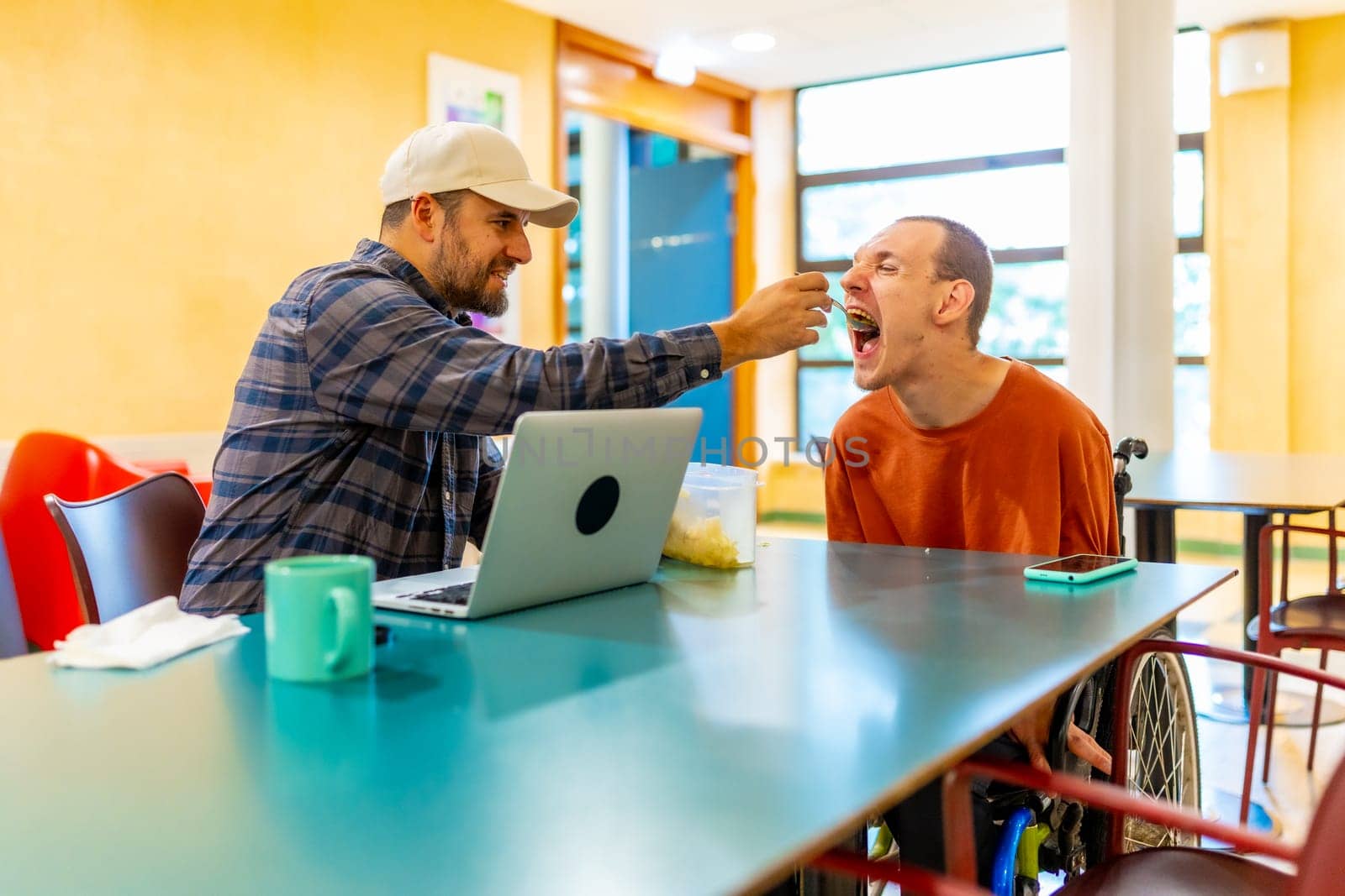 Man feeding a coworker with disability in the public cafeteria of a modern coworking