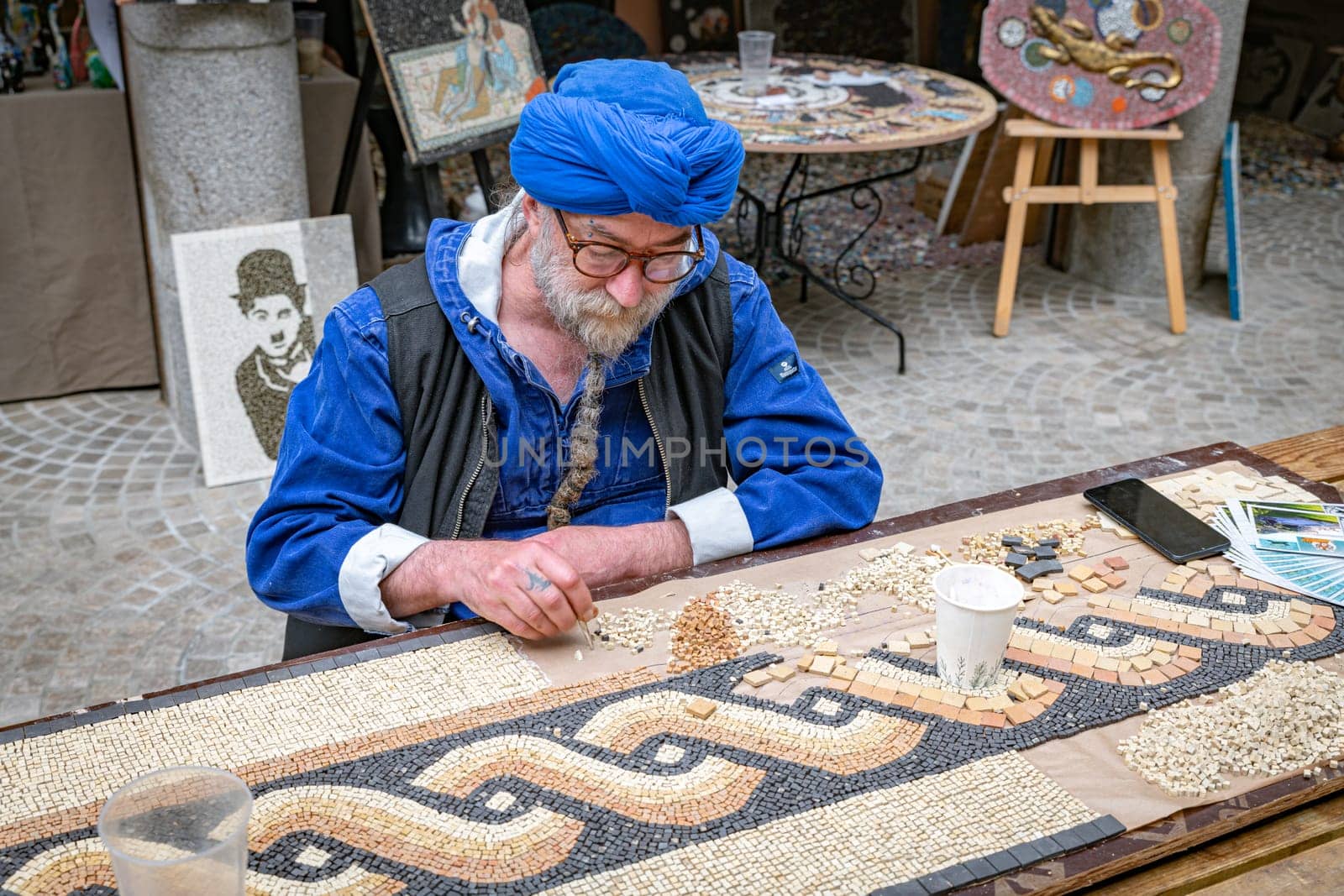 dinan,france,28-04-2024:a man sits at a table and makes art by putting pieces of stone together like mosaics to create beautiful images like paintings