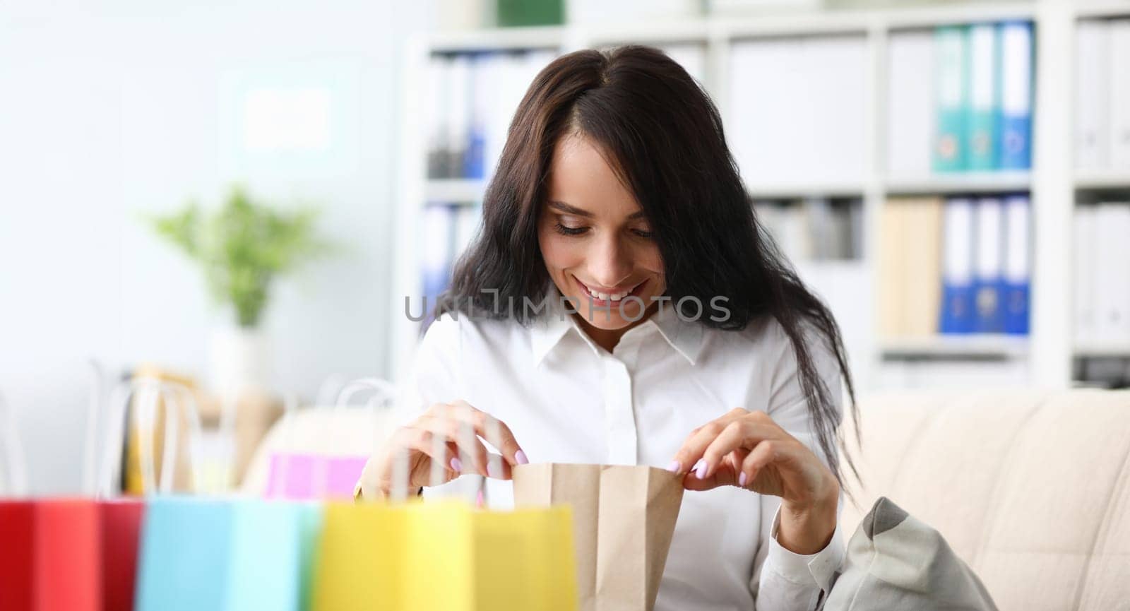 Portrait of pretty businesswoman looking in multicolored shopping packets. Brunette sitting in modern office on comfortable sofa and looking for purchases. Shopping sale concept