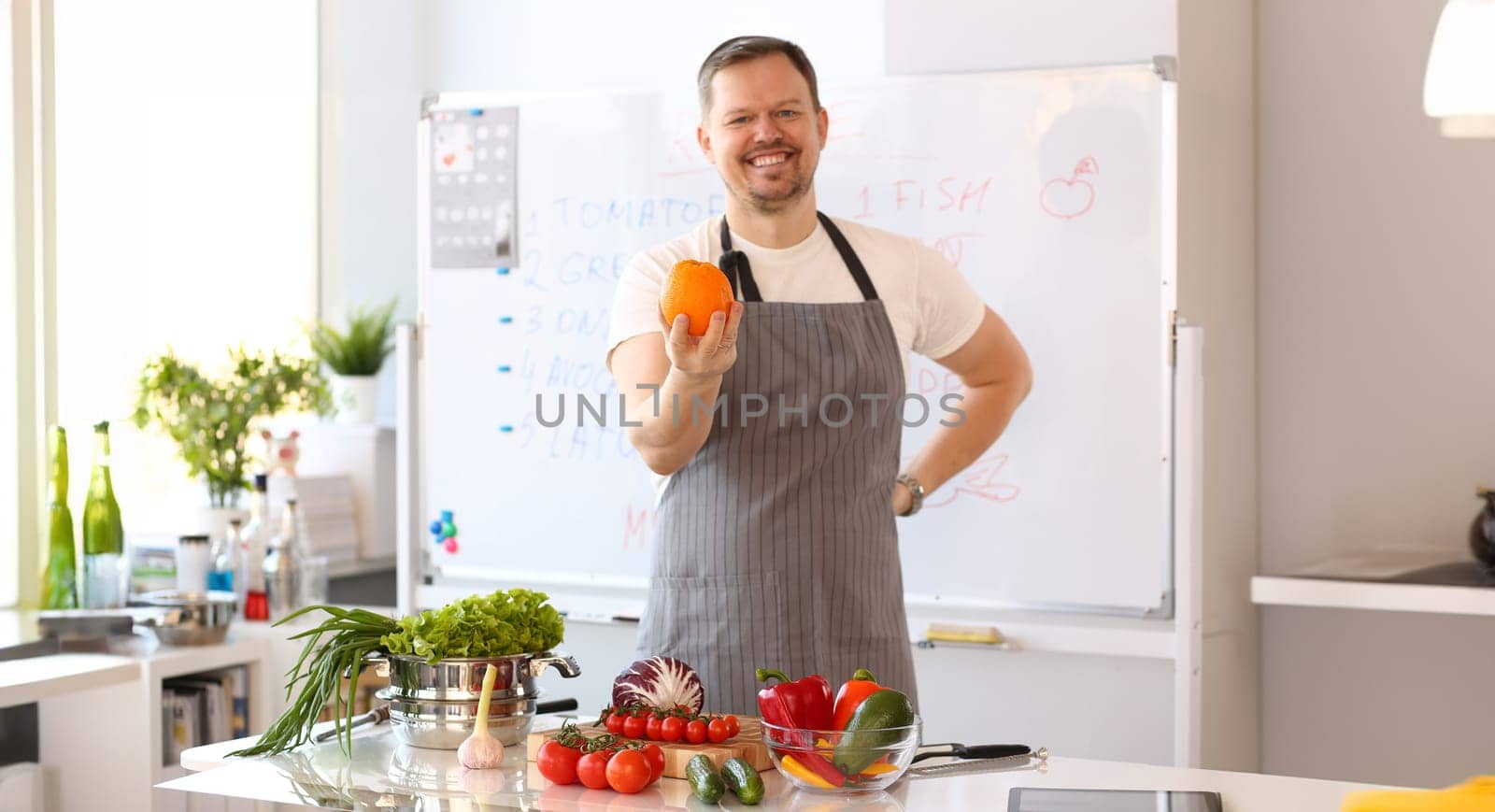 Man Blogger Holding Orange Cooking Vegetables Portrait. Male Cook Salad at Home in Kitchen. Culinary Recipe from Tomatoes, Cucumber, Cabbage and Garlic. Smiling Chef in Apron Looking at Camera Shot