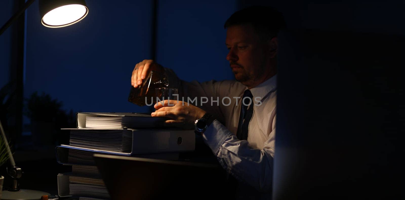 Portrait of tired businessman sitting indoors and pouring alcohol in glass. Man relaxing after intense workday with pile of paper folders full of corporation documents. Night at office concept