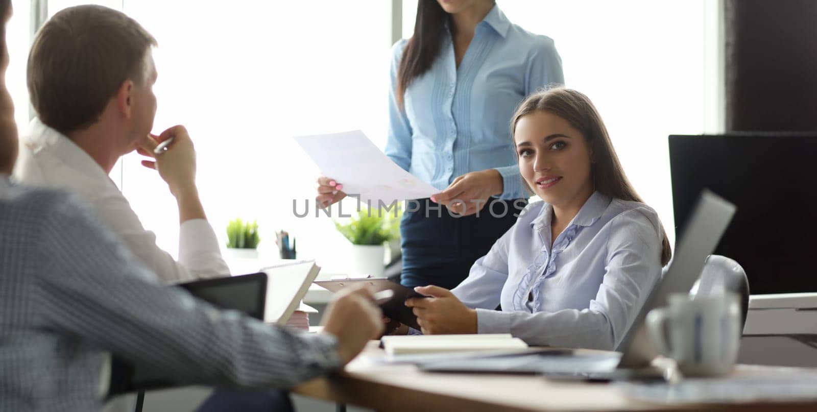 Portrait of positive woman sitting at table in office. Businesslady in classic shirt and looking at camera with calmness and gladness. Team discussing important documents with boss. Teamwork concept