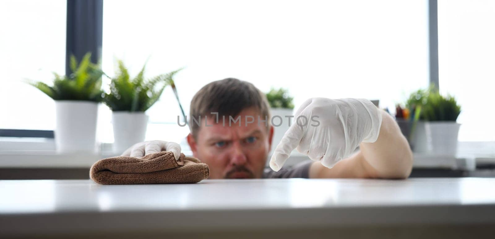 Focus on gloved hand of diligent male touching white office workplace and holding brown loincloth and swiping dust with harmful germs. Pleasant clean-up concept. Blurred background