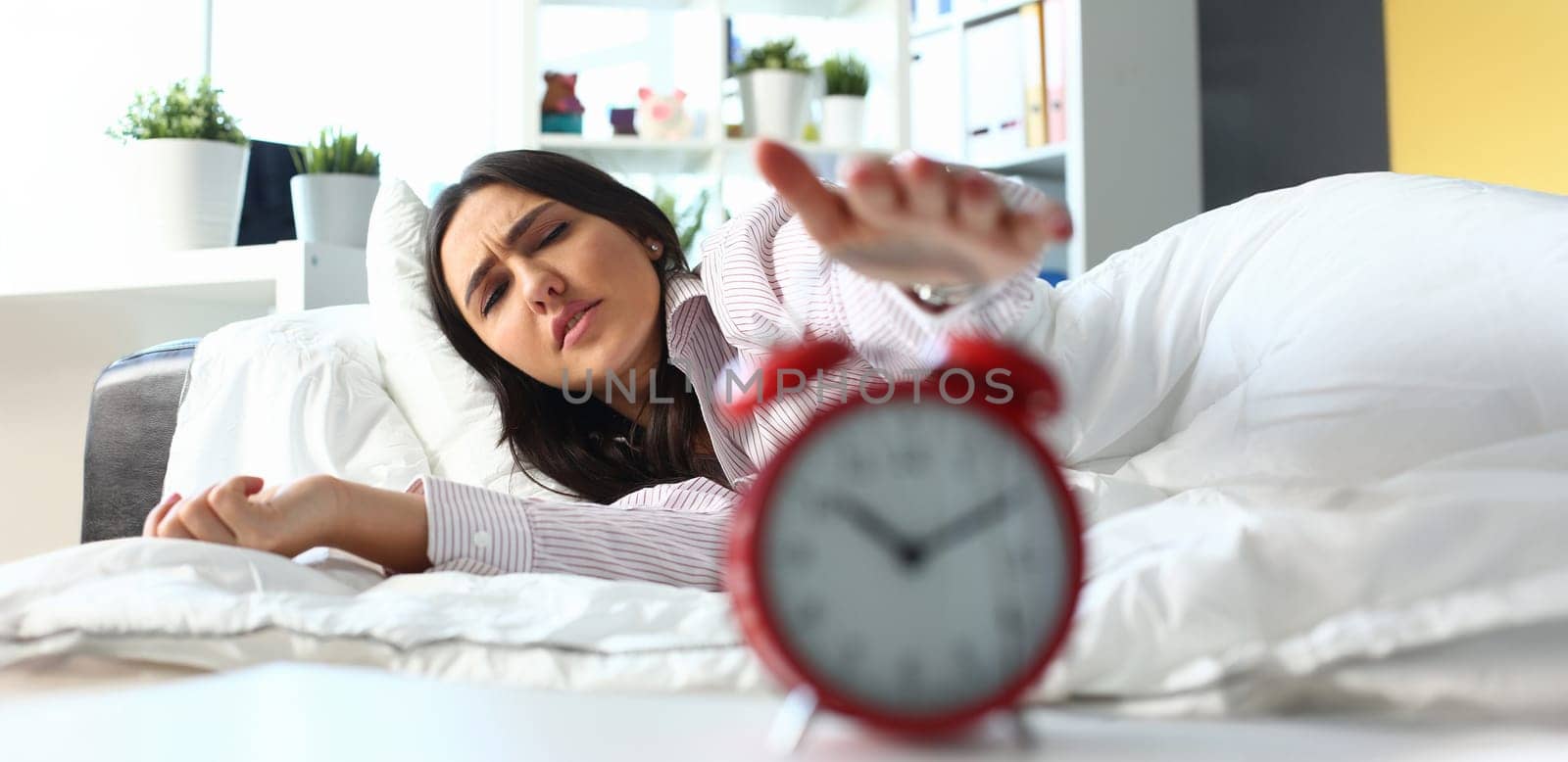 Sleepy young woman portrait with one opened eye trying kill alarm clock. Early wake up not getting enough sleep going work concept. Female stretching hand to ringing alarm willing turn it off