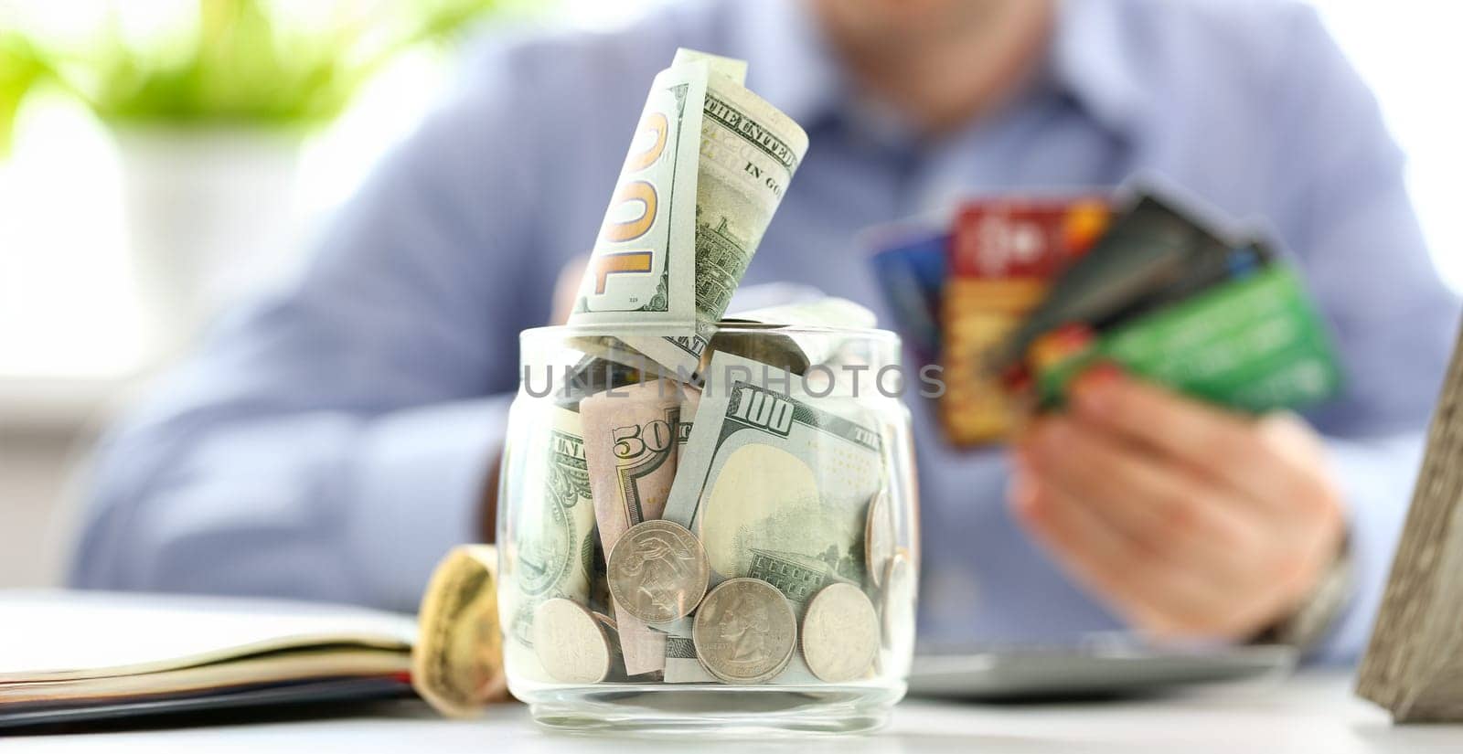 Big jar full of money stand at working table with male hand holding bunch of credit cards in background