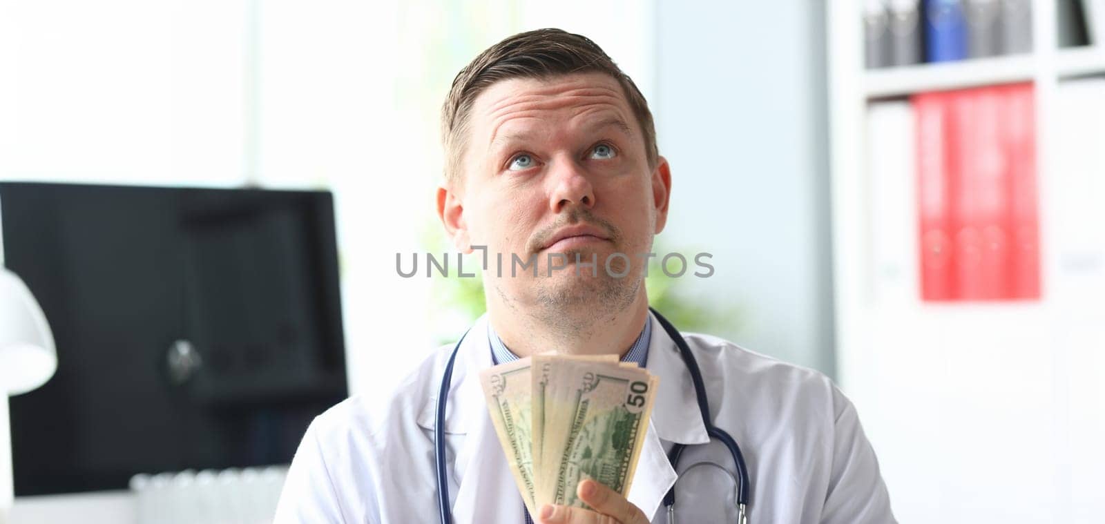Portrait of smart professional physician in modern hospital office holding fan of green banknotes and looking up with satisfaction. Healthcare and clinic concept