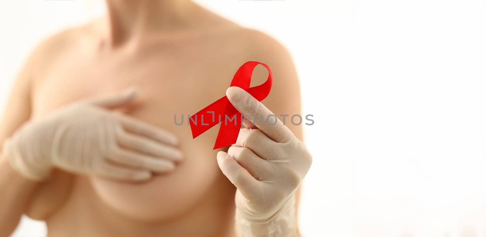 Female hand in gloves holding red ribbon cancer symbol breast closeup on doctor mammology clinic reception. Mammary glands test biopsy implant silicone insert tumor human concept