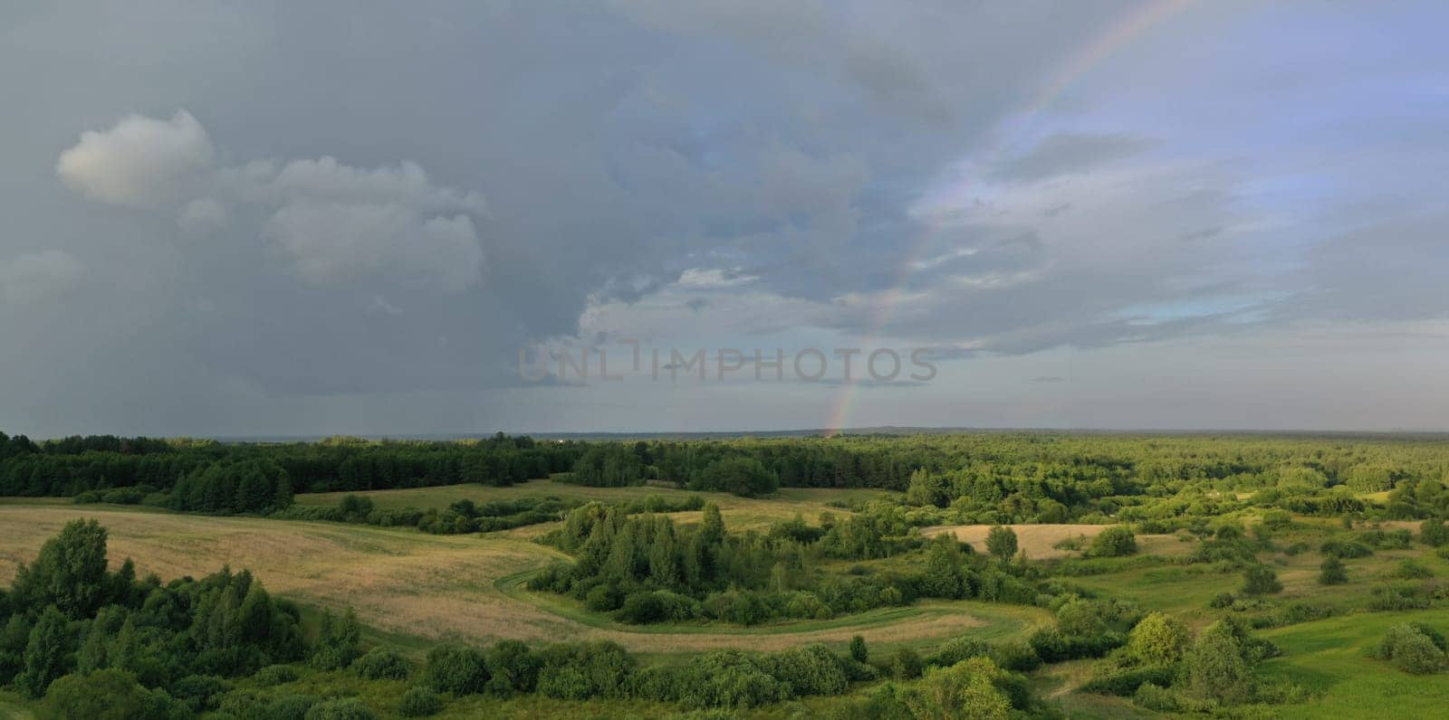 Top view of meadow with road and green plants. Cloudy sky and beautiful rainbow above trees and bushes. Picturesque landscape scene of rain in summer time