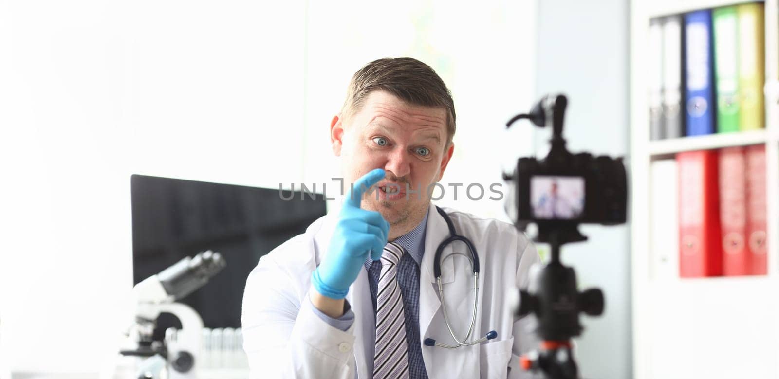 Portrait of smart physician sitting at modern hospital workplace and showing one finger causing too much inconvenience to high-tech camera. Healthcare and clinic concept