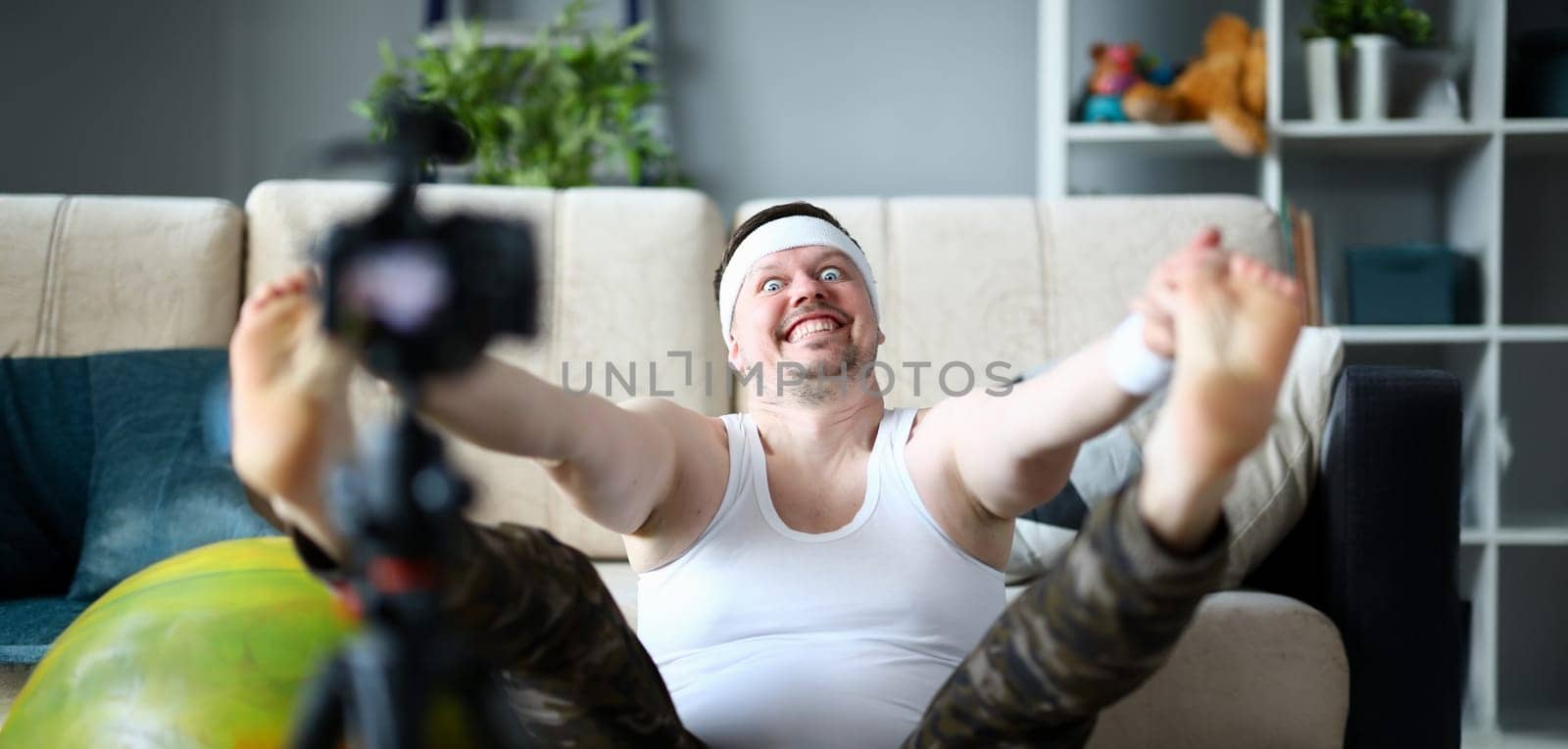 Vlogger freak shows his subscribers on camera how not to do the stretching correctly, holding his hands over his feet. Astonished face expression grimaces aganist home background concept