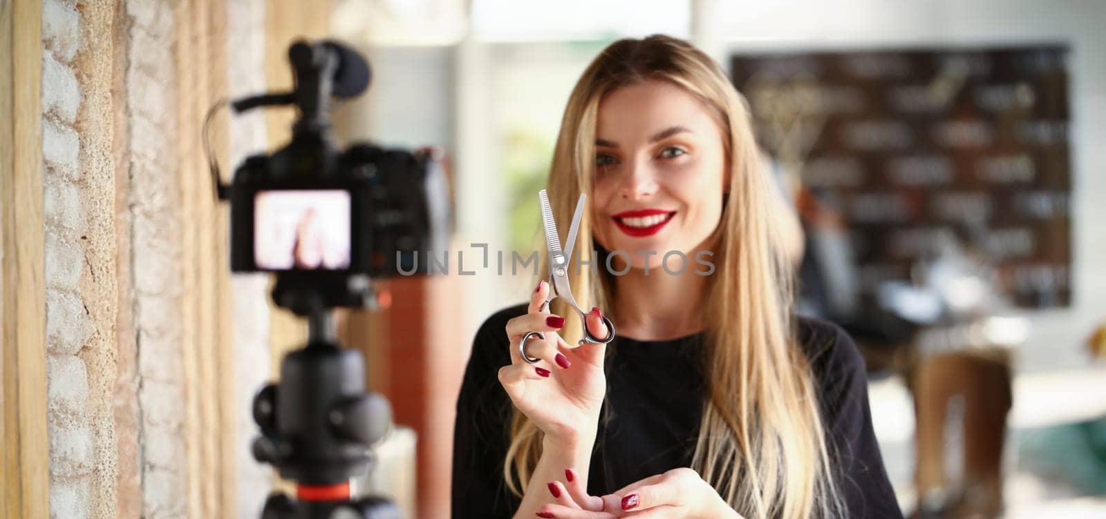 Female millennial vlogger hold in hand scissors aganist barbershop background. Study how to make fashionable modern concept haircuts.