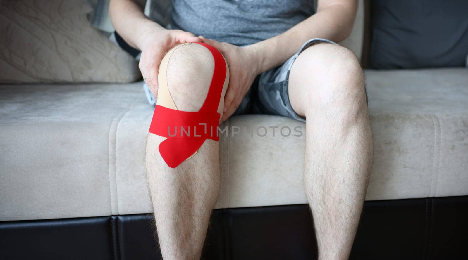 Male hands holding sored knee sitting on couch at home closeup