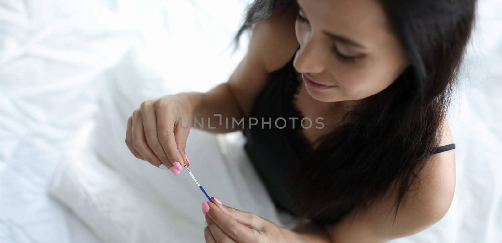 A lonely woman sits on bed and holds pregnancy test in her hand. Female maternity reverso context concept.