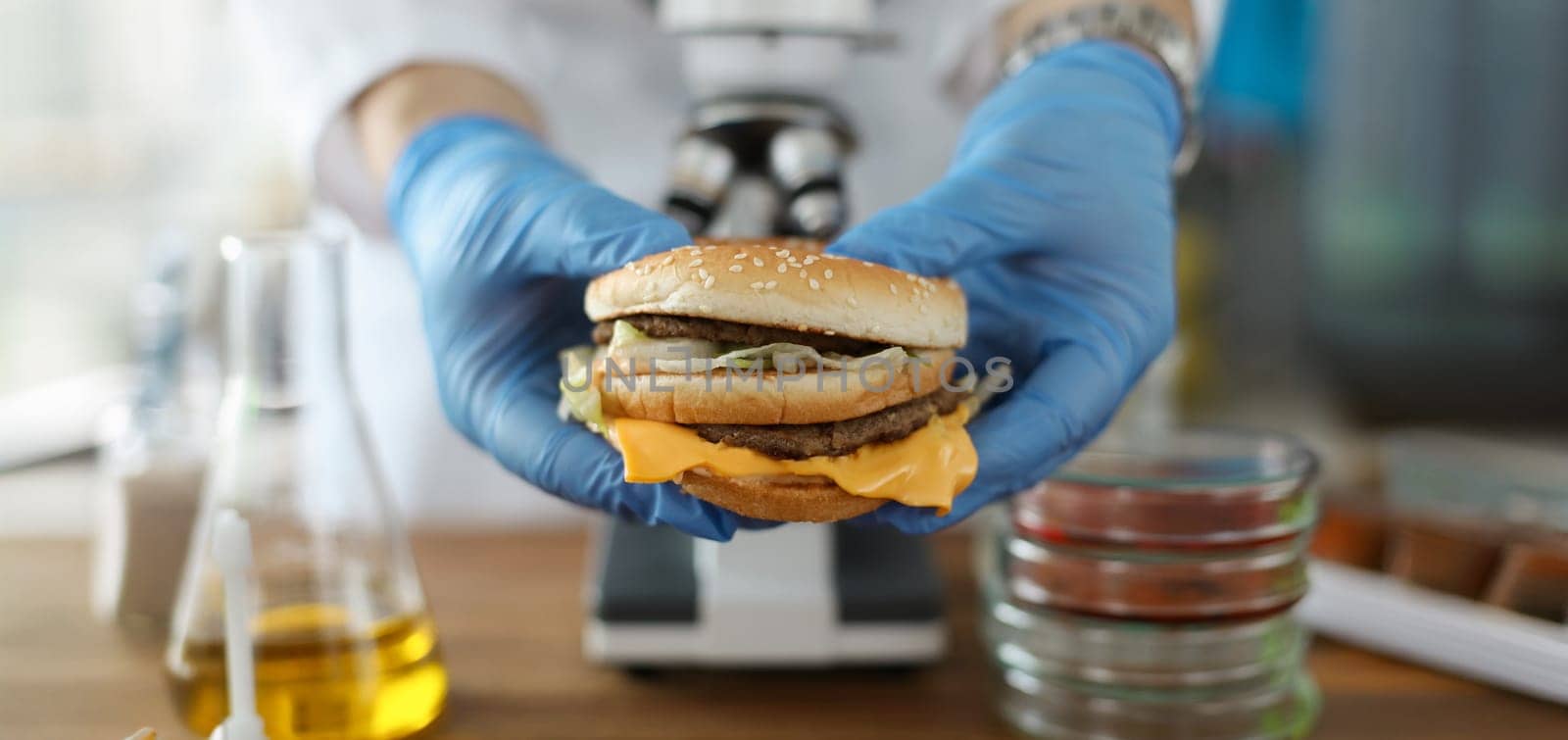 Male hand hold tasty burger in hand with blue protective gloves closeup. Testing food nutrition concept.