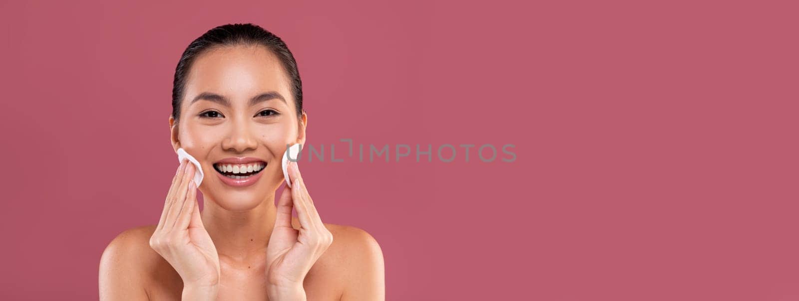 Closeup photo of happy topless young korean woman cleansing her skin with cotton pads and smiling over purple studio background, removing makeup with beauty product, copy space