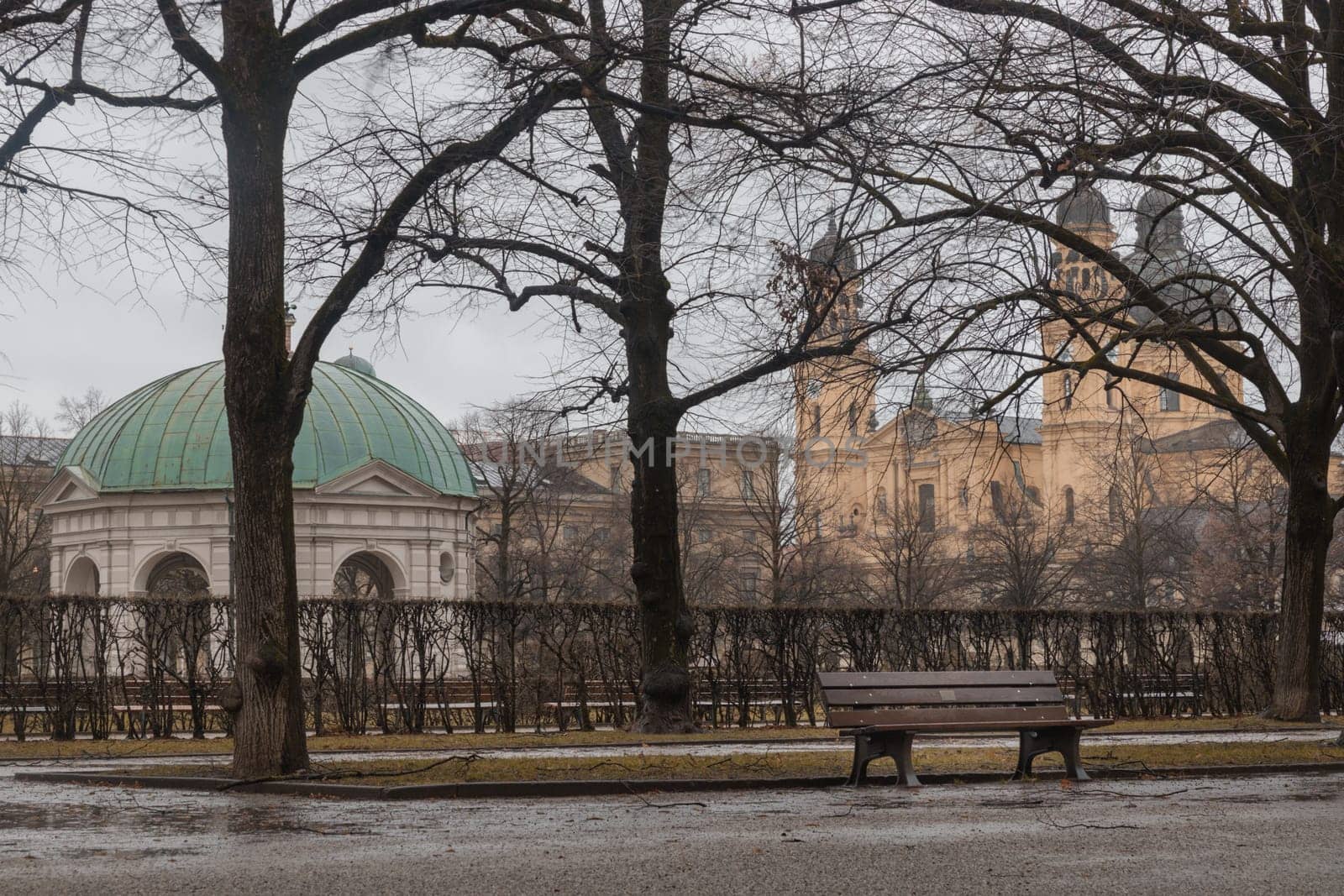 Munich, Germany - Dec 21, 2023 - Bench made wood and iron stand in the garden of the Munich Residenz with The Diana Temple (Dianatempel) and St.Cajetan and Adelaide in the background. Space for text, Selective focus.
