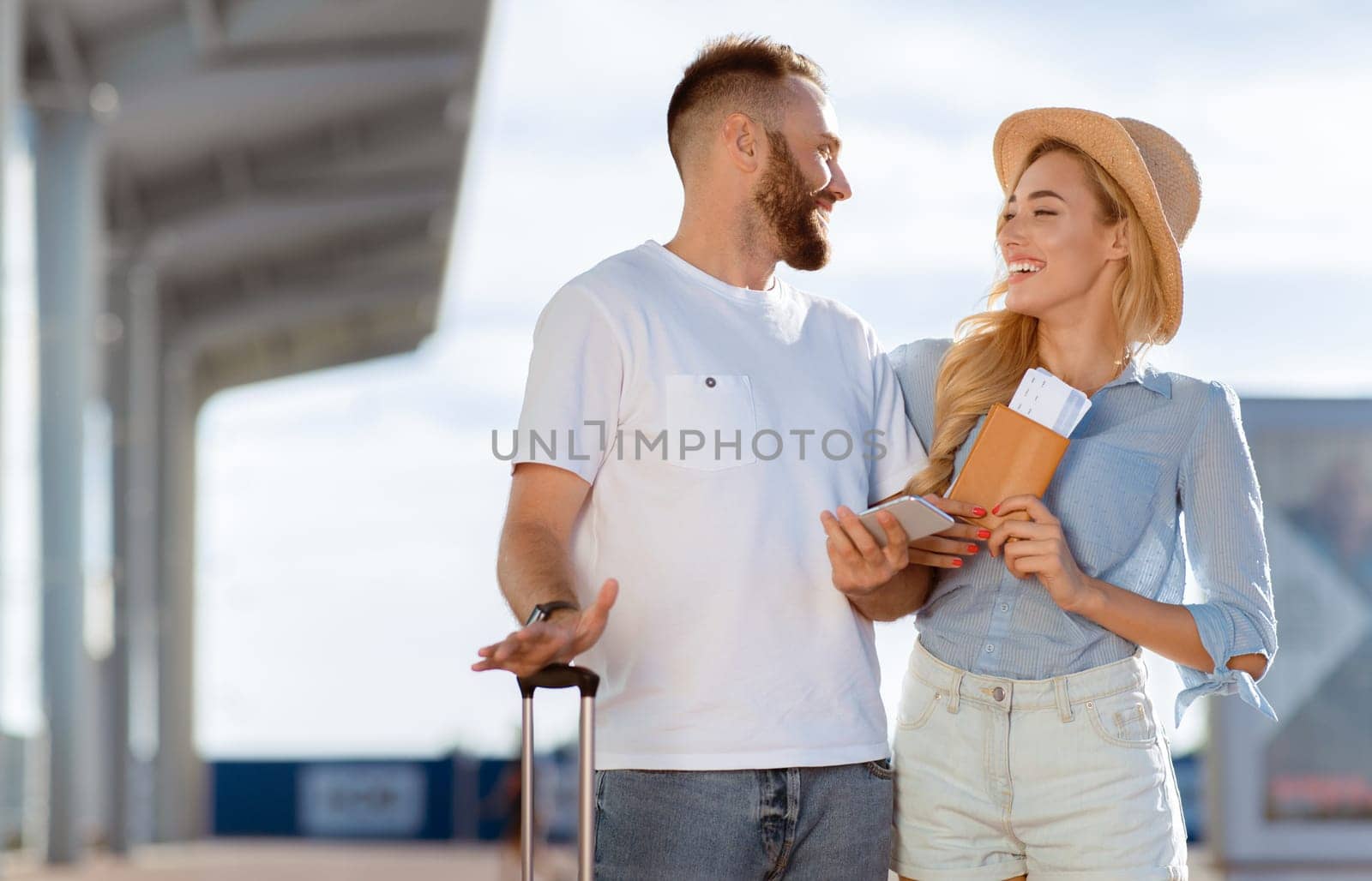 Vacation Concept. Cheerful casual couple standing with valise outside, looking at each other, copy space