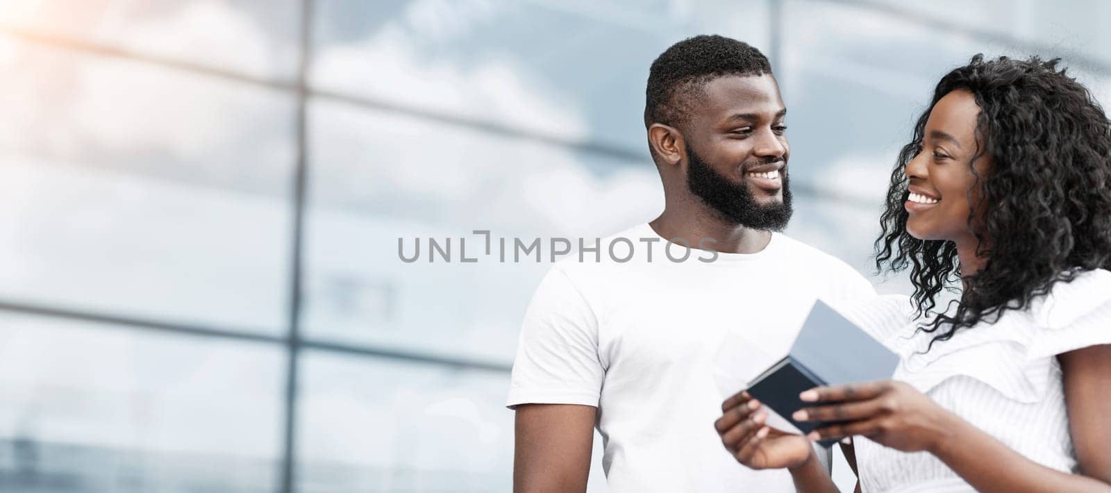 Black man and woman, dressed in casual clothing, stand outdoors and smile at each other while looking at documents. The couple is likely preparing for a trip, copy space
