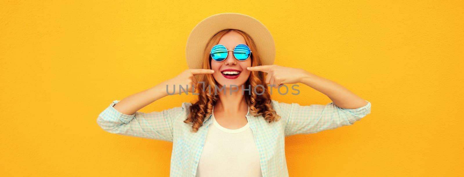 Portrait of stylish happy smiling young woman points with fingers to her white clean teeth in summer hat, glasses on vivid yellow studio background