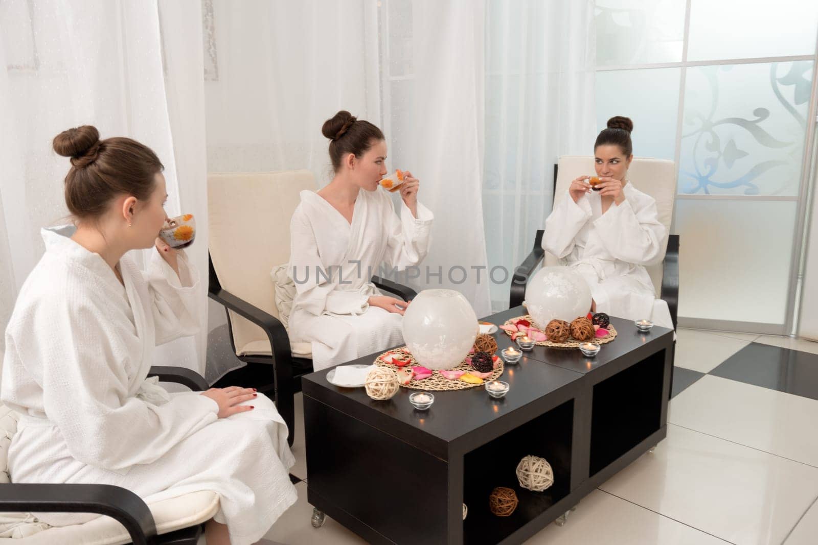 Three pretty young girls with nice bundles in white bathrobes sitting by the table laid for tea and relaxing shot in spa saloon
