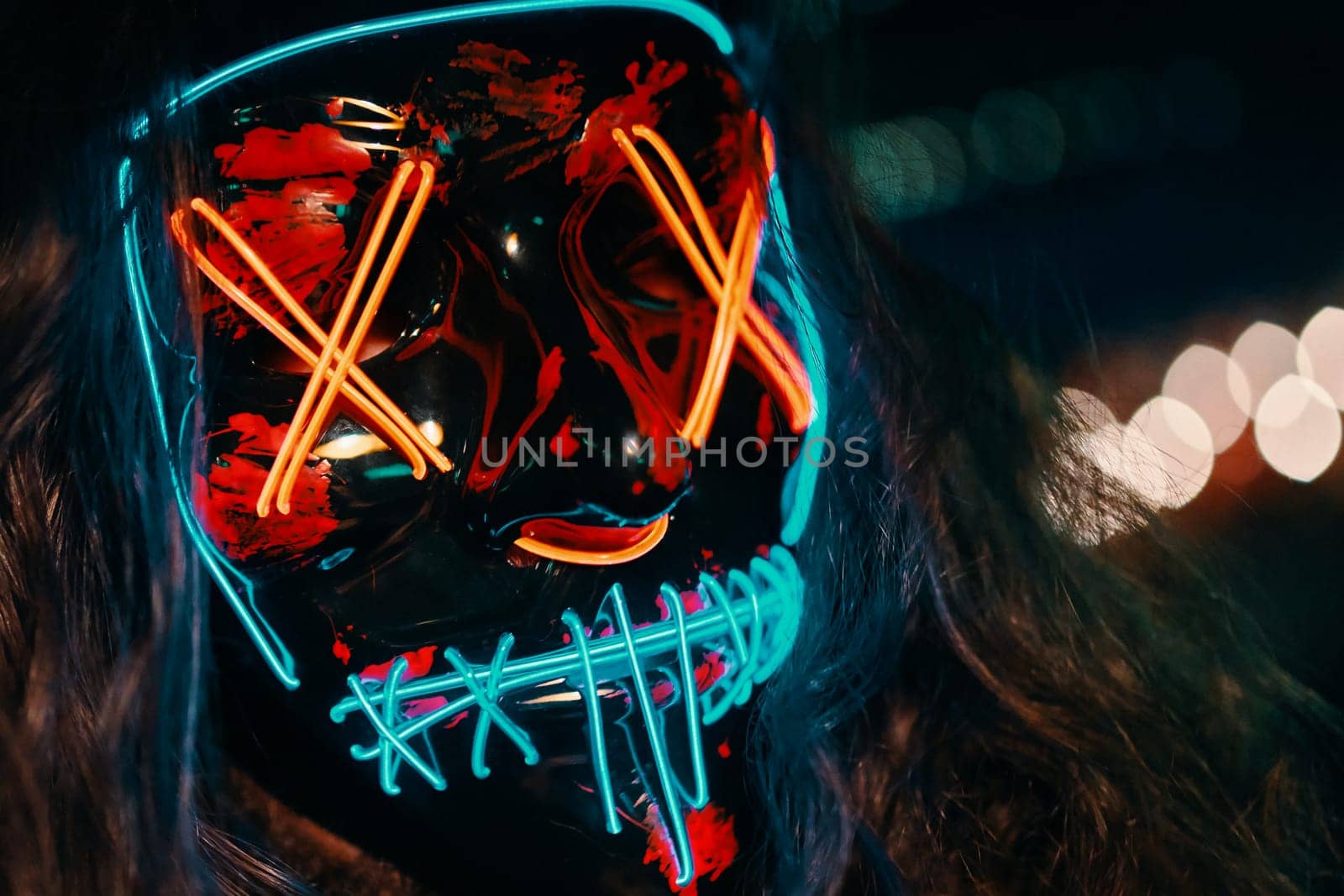 Close up of man with long disheveled hair in a lighting neon glowing mask. Halloween dress-up party in night club. Anonymous person is a cyber criminal. All Saints' Night.