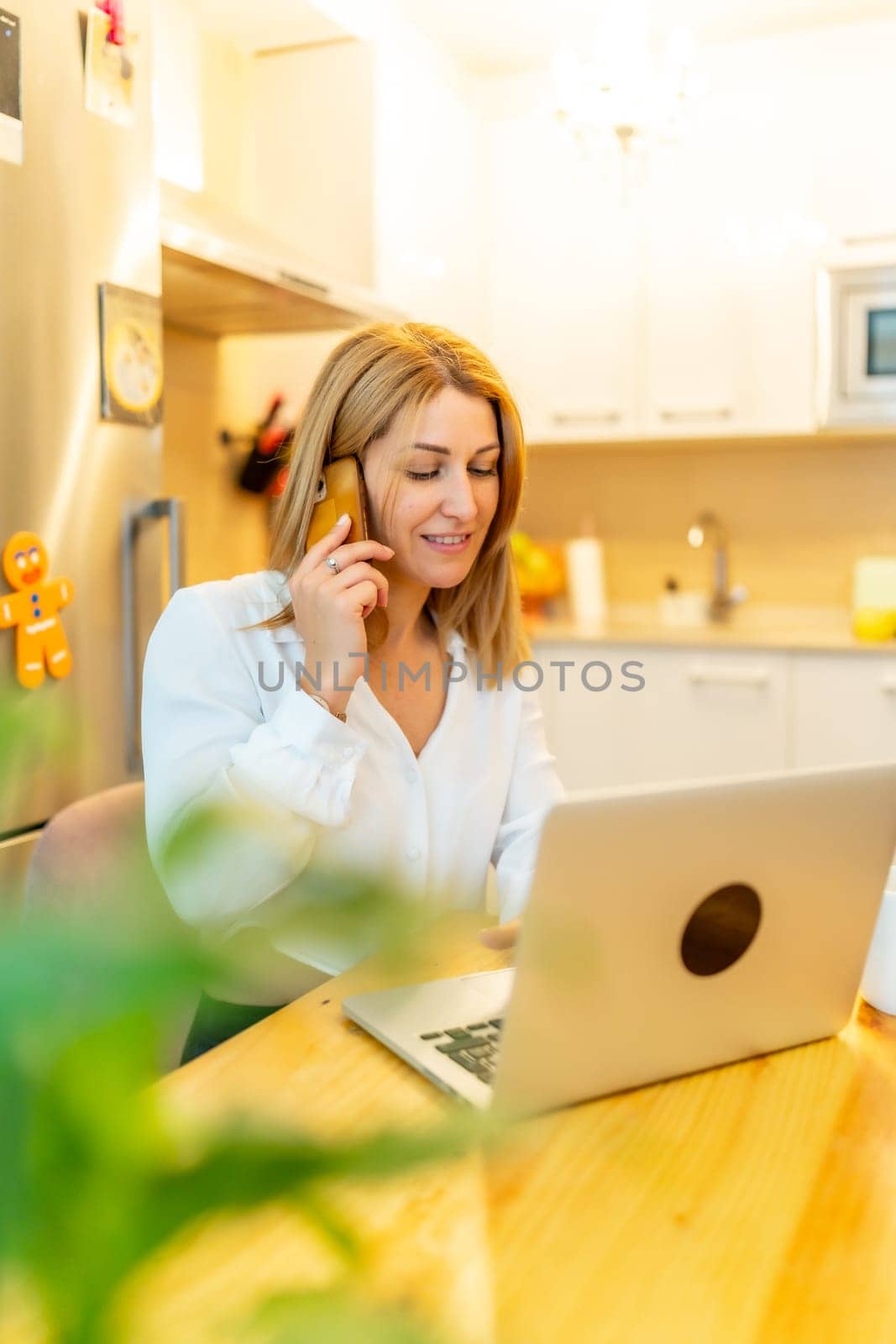 Vertical photo of a blonde businesswoman attending a call working from home sitting on a table next to the kitchen