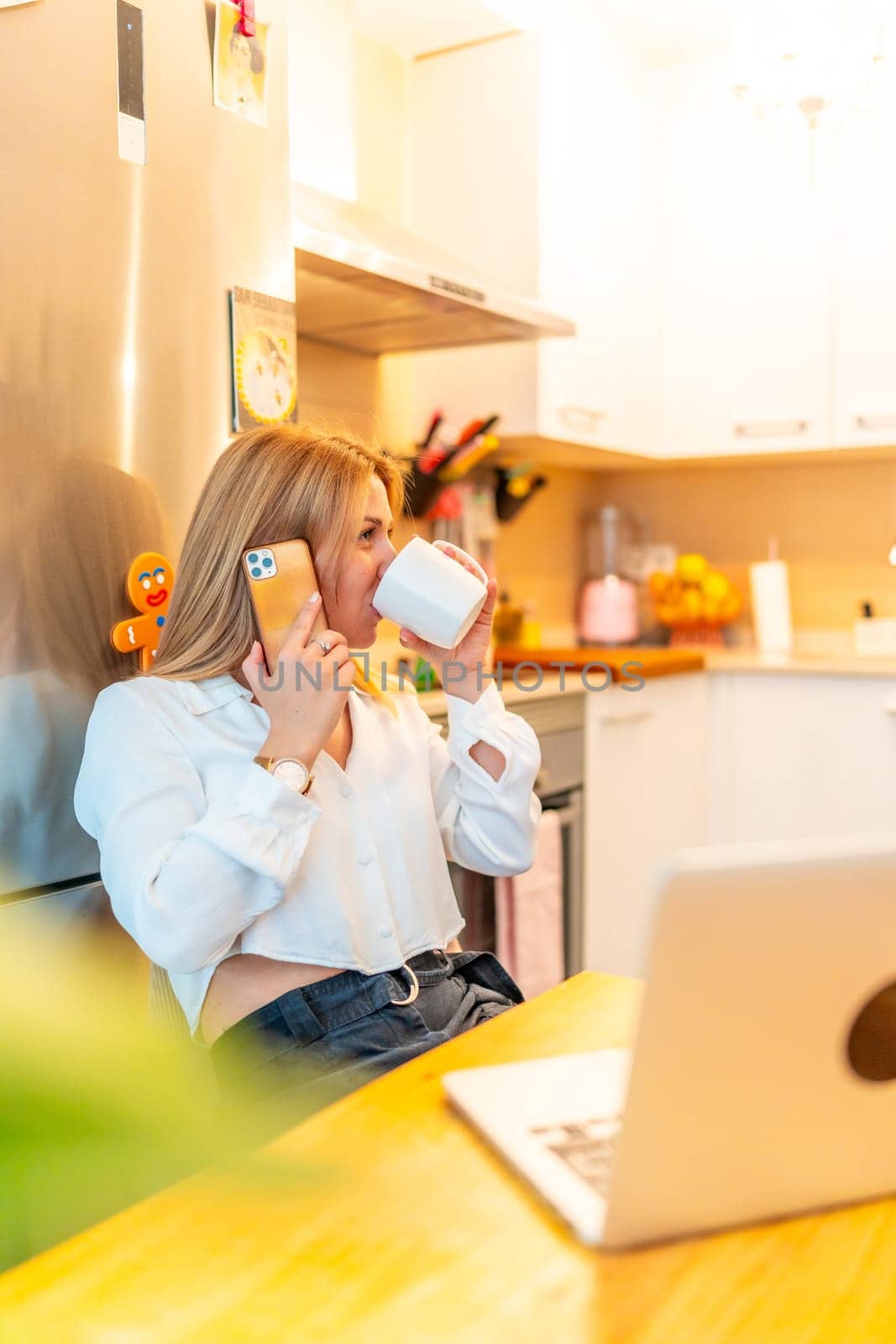 Vertical photo of a blonde caucasian female freelancer drinking coffee while working from home using mobile phone and laptop