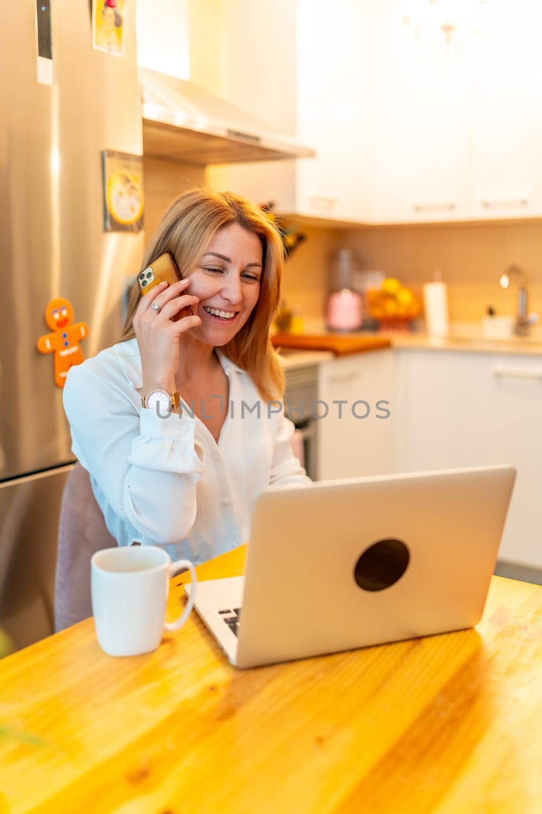 Vertical photo of a blonde caucasian adult businesswoman working from home in the morning using laptop and mobile phone next to coffee cup