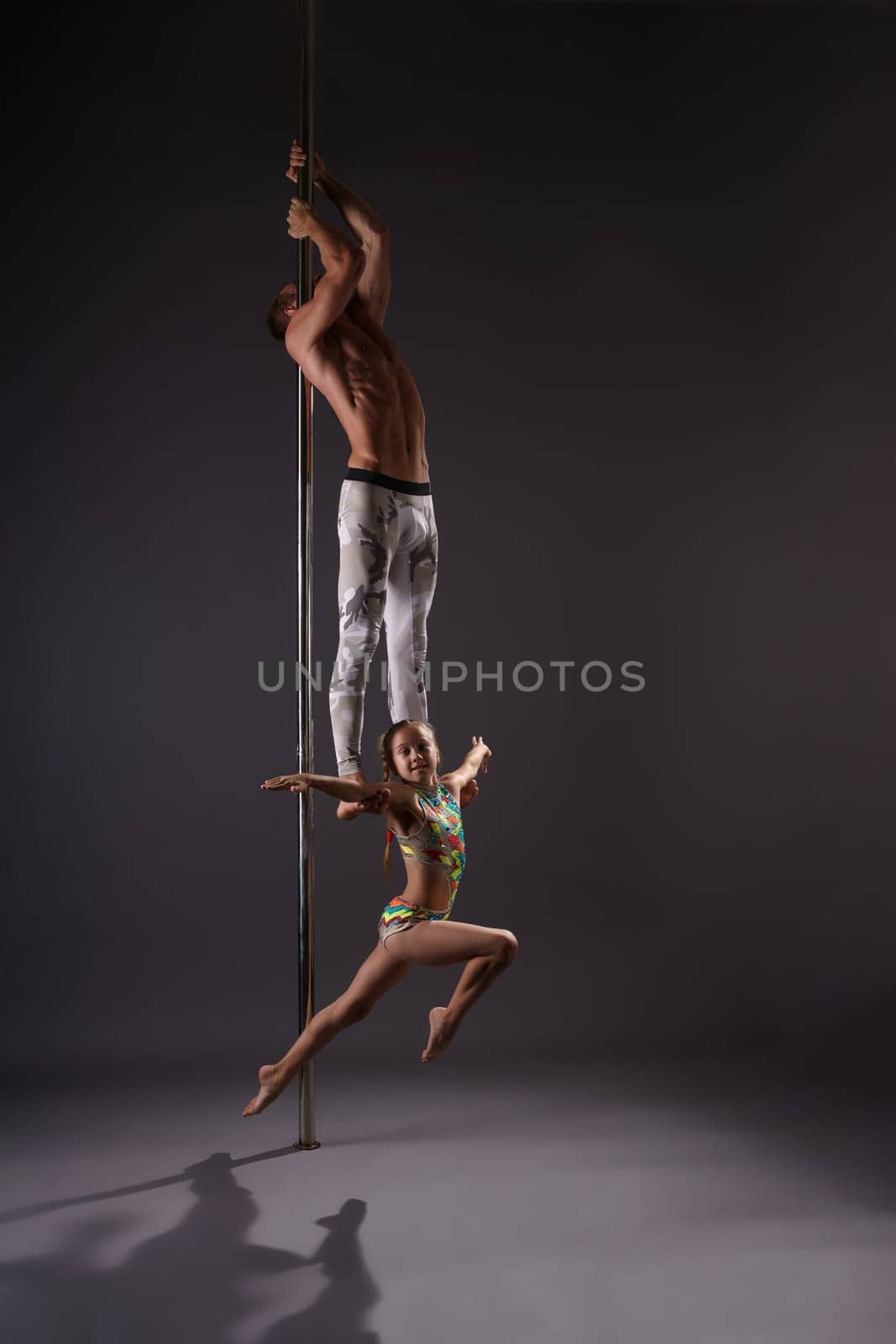 Athletic young male pole dancer making exercises in pair with little girl in studio