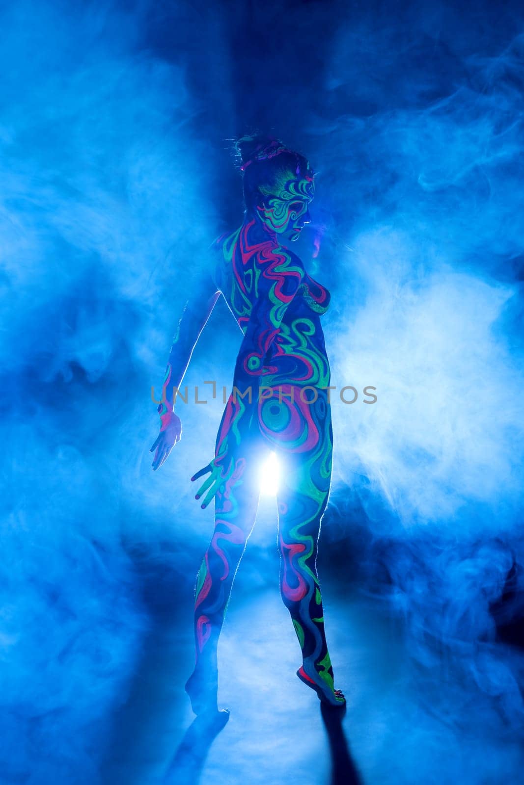 Slim girl posing in smoky studio her face and figure covered by bright fantastic ultraviolet color pattern bodyart.