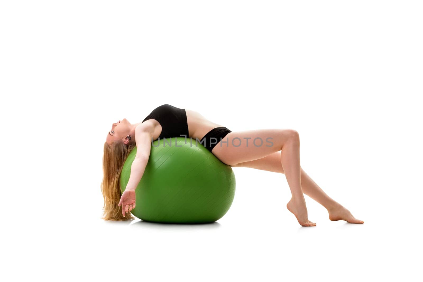 Long haired blonde in black shorts and briefs lying on her back on green big fitball posing in studio