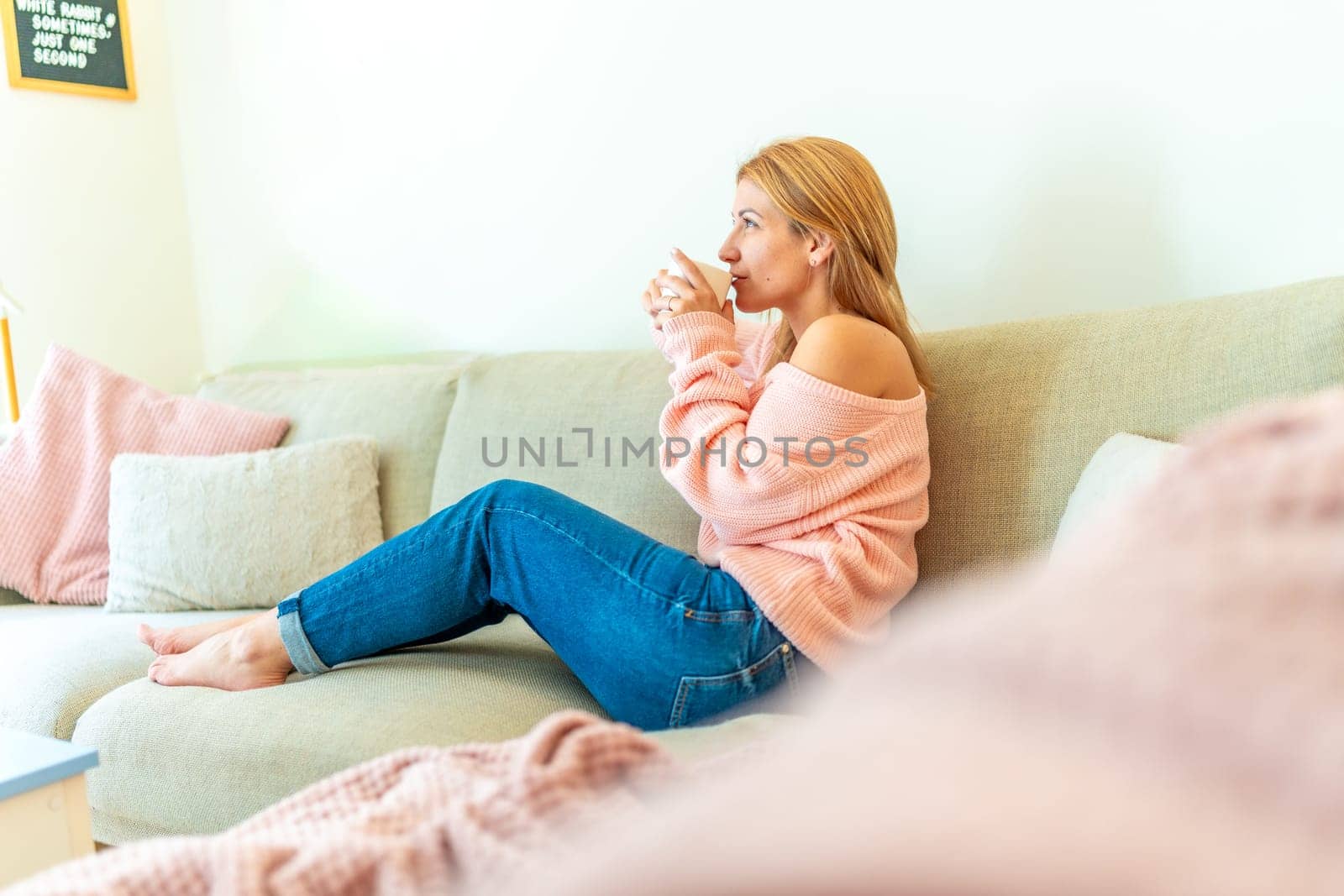 Full length side view of a blonde caucasian adult relaxed woman sipping coffee sitting comfortable in the sofa