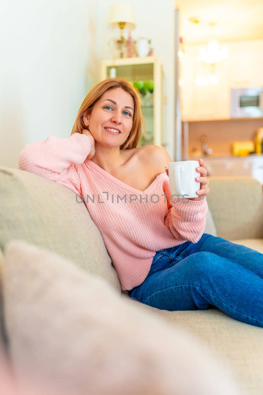 Vertical photo of a mature blonde beauty woman relaxed on the sofa sipping coffee