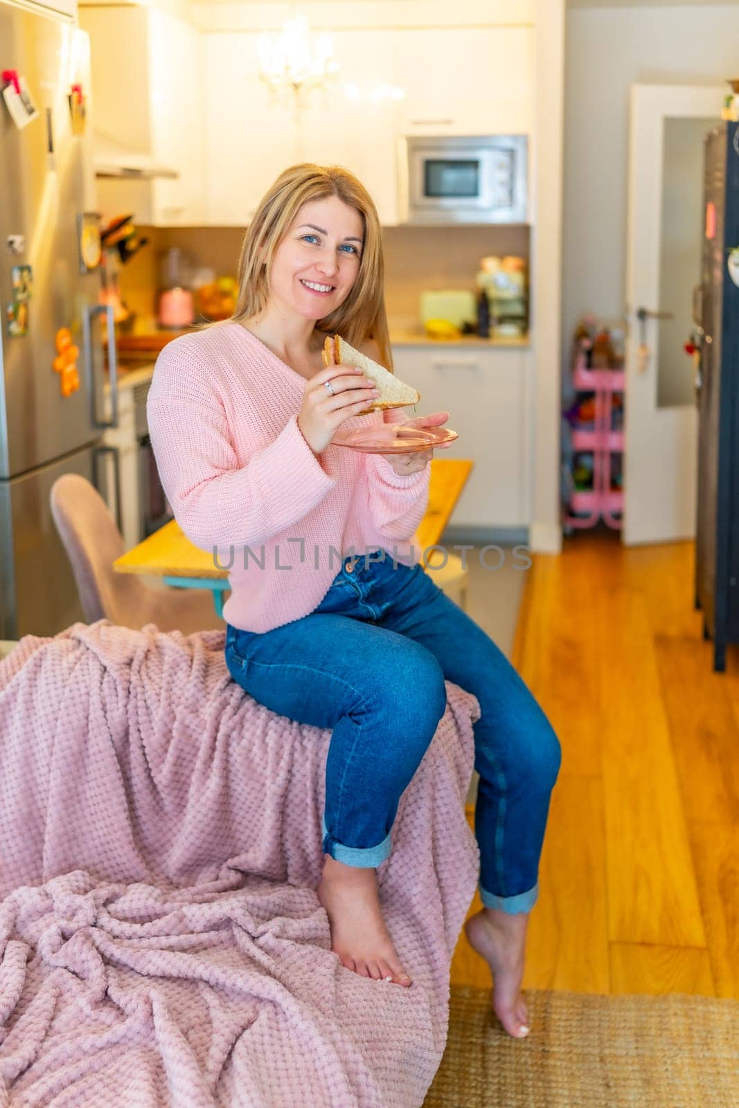 Vertical portrait of a mature adult caucasian blonde smiling comfortable woman eating healthy sandwich at home