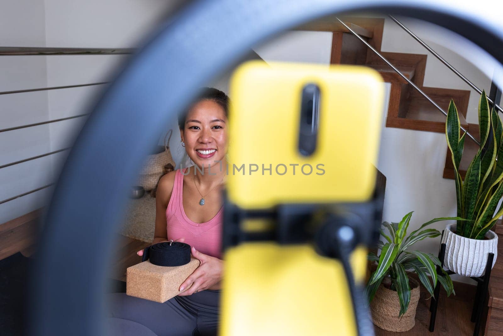 Focus on background. Happy young asian woman recording yoga class for social media using mobile phone and tripod. by Hoverstock