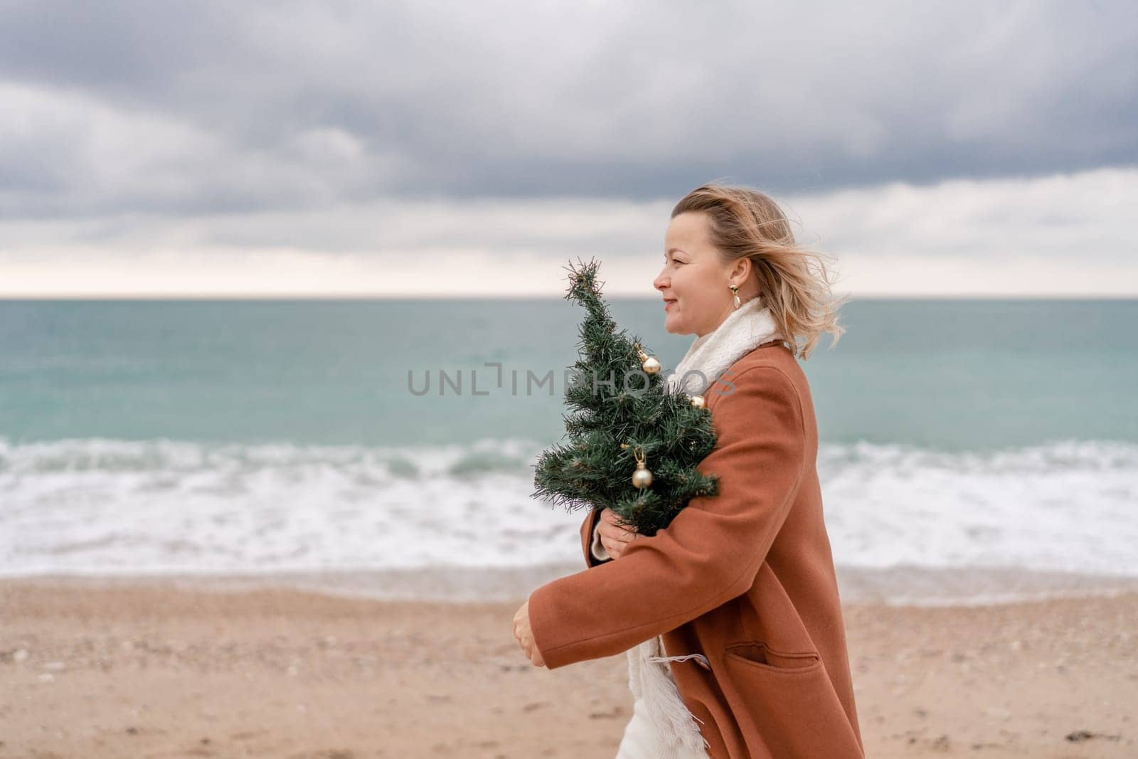 Blond woman Christmas sea. Christmas portrait of a happy woman walking along the beach and holding a Christmas tree on her shoulder. She is wearing a brown coat and a white suit