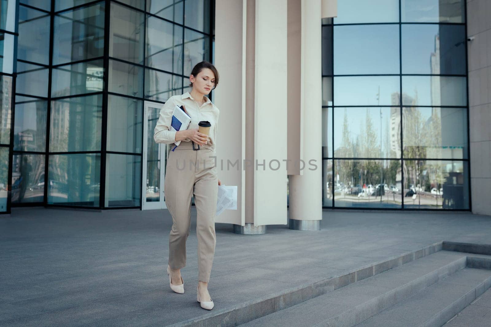 Business woman hurries out of an office building with coffee and documents in her hands. Time Management Issues.