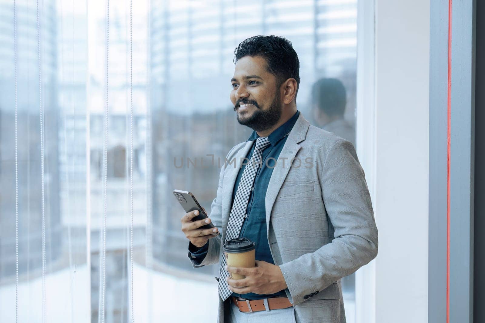 Indian businessman standing next to window using mobile phone wile coffee break by itchaznong