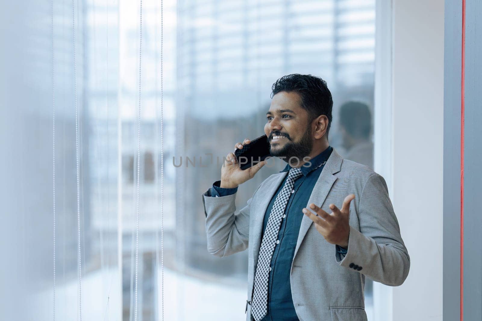 Indian businessman using smartphone talking having phone call conversation in office looking out window.